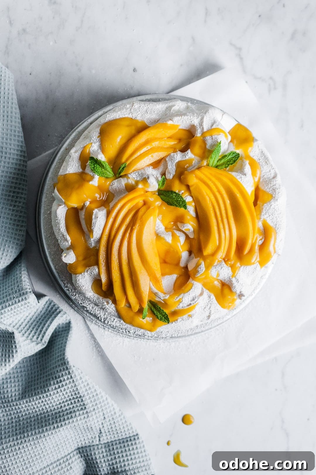 Top view of meringue cake with mango sauce and sliced mangoes on top, garnished with mint leaves on a white marble background