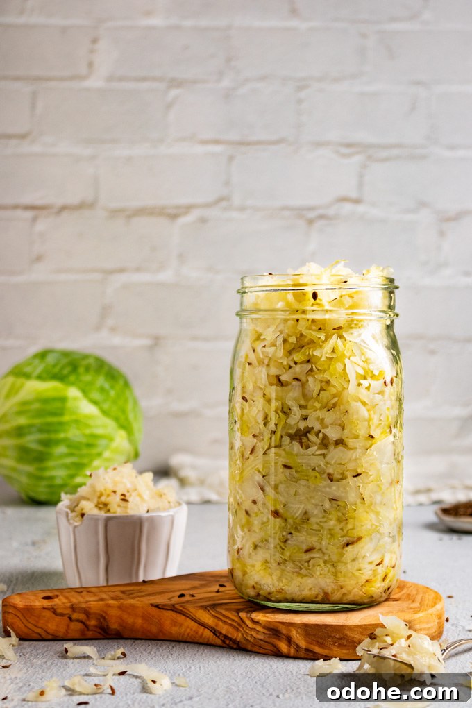 Straight on shot of homemade sauerkraut in a large mason jar. The jar is on a wooden board, there is a small fork with more sauerkraut laying next to the jar, a small bowl of sauerkraut is off the the side, and a head of green cabbage is in the background. The backdrop is white brick.