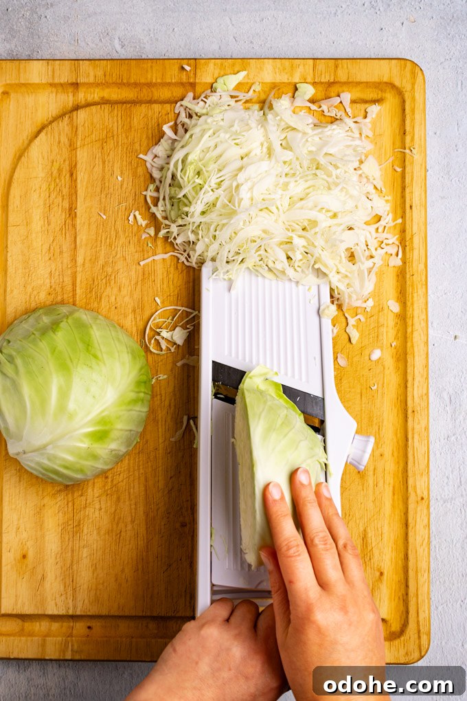 Slicing a head of cabbage on a mandolin. A hand is holding a piece of cabbage, and shredded cabbage is piled at the end of the mandolin.