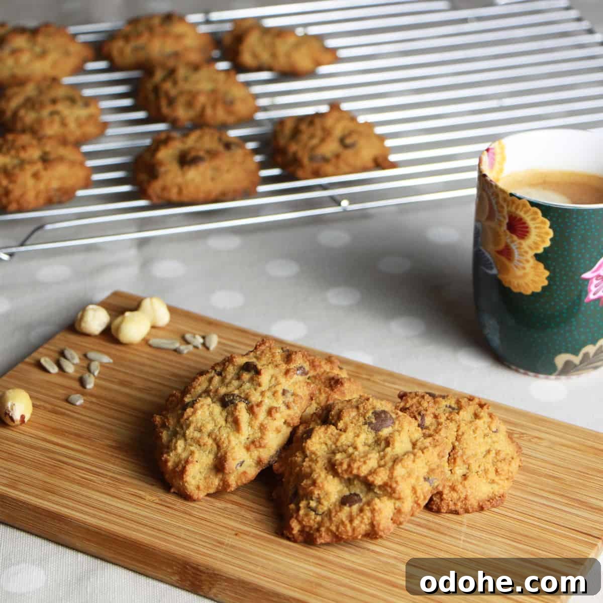 Photo of a chopping board with hazelnut choc-chip cookies on it and cup of coffee in the background with a cooking rack of more cookies behind.