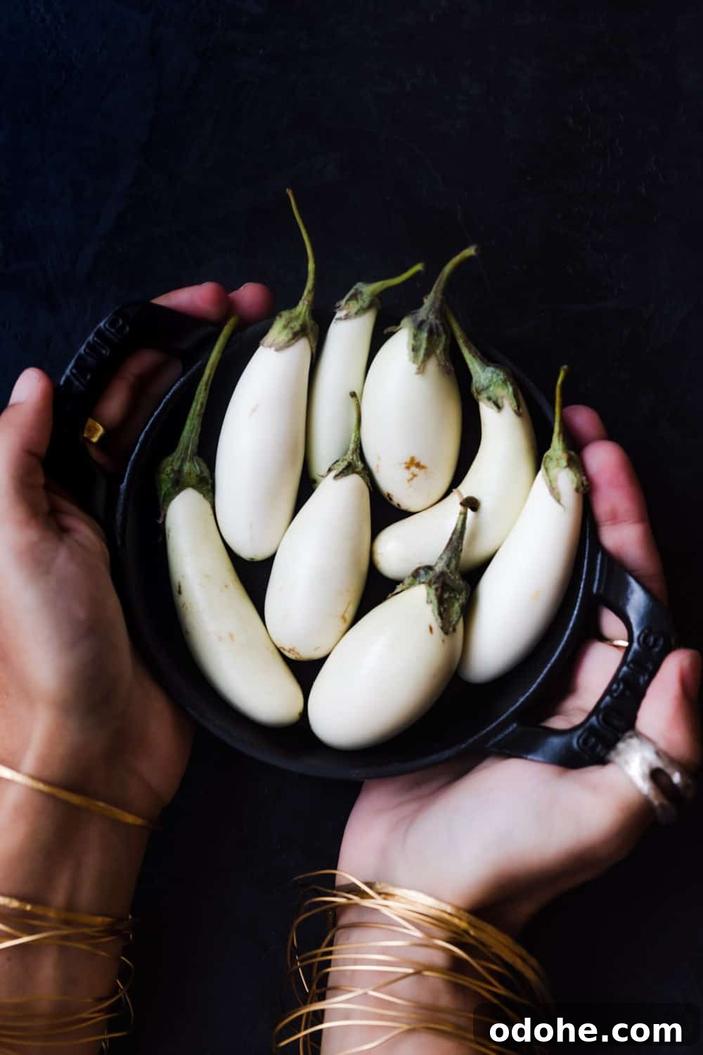 Hands holding a mini black skillet filled with small white eggplants against a dark background.