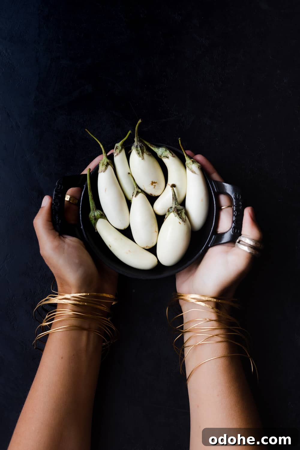 Hands holding a black mini skillet filled with small white eggplants against a dark background.