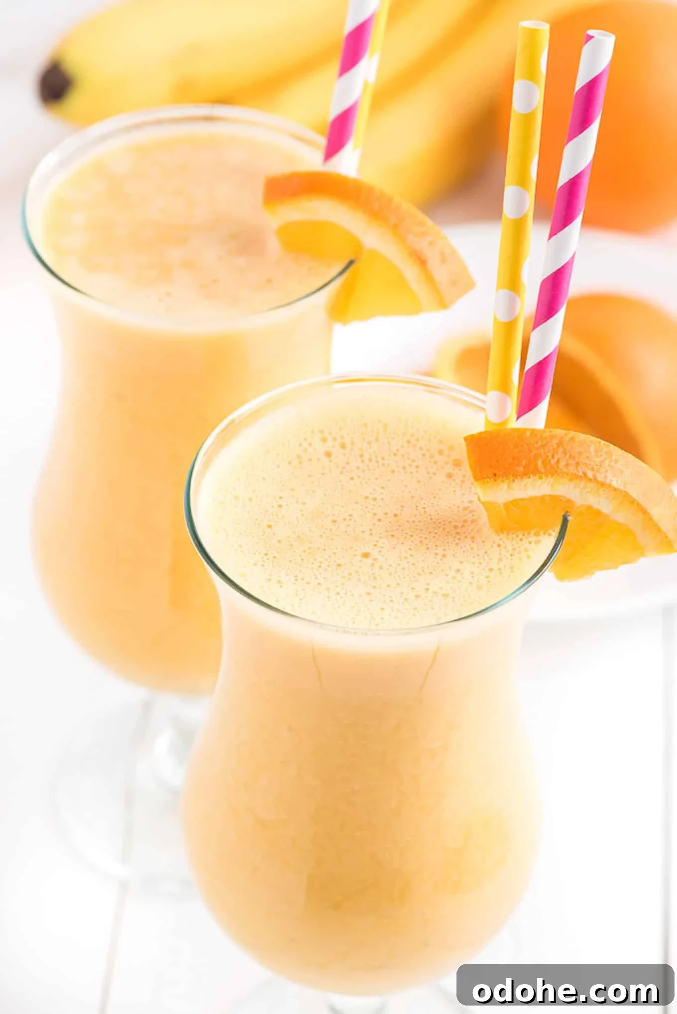Energizing Orange Smoothie Recipe for Vitamin C Boost 2 Tall glass of orange smoothie with an orange slice on the side and a pink straw.