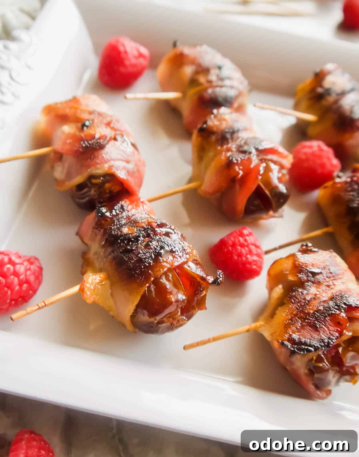 Bacon-Wrapped Raspberry-Stuffed Dates | Paleo & Whole30-Friendly 2 Raspberry stuffed Dates Wrapped in Bacon on a plate with raspberries surrounding them.