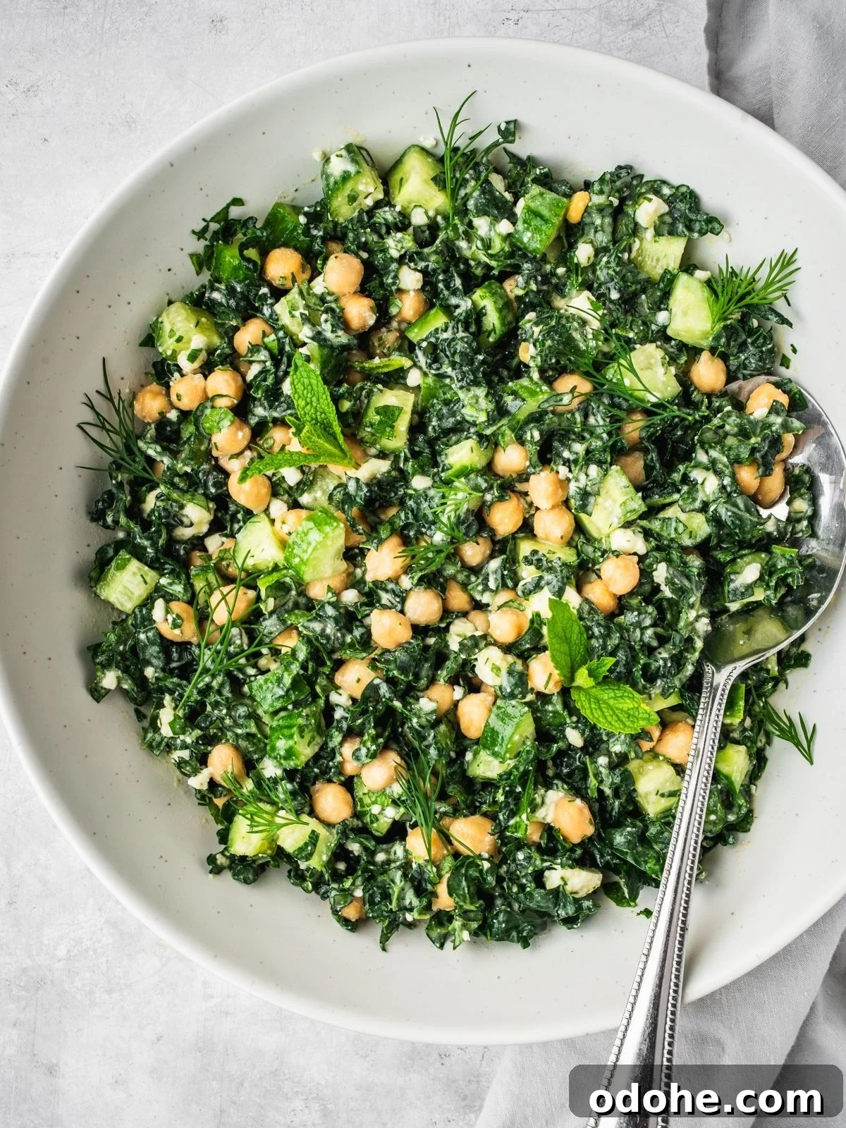 A bowl filled with a fresh salad made of kale, chickpeas, cucumber, herbs, and crumbled cheese. A spoon rests in the bowl.
