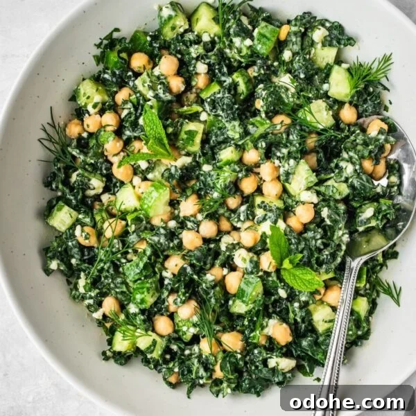 A bowl filled with a fresh salad made of kale, chickpeas, cucumber, herbs, and crumbled cheese. A spoon rests in the bowl.