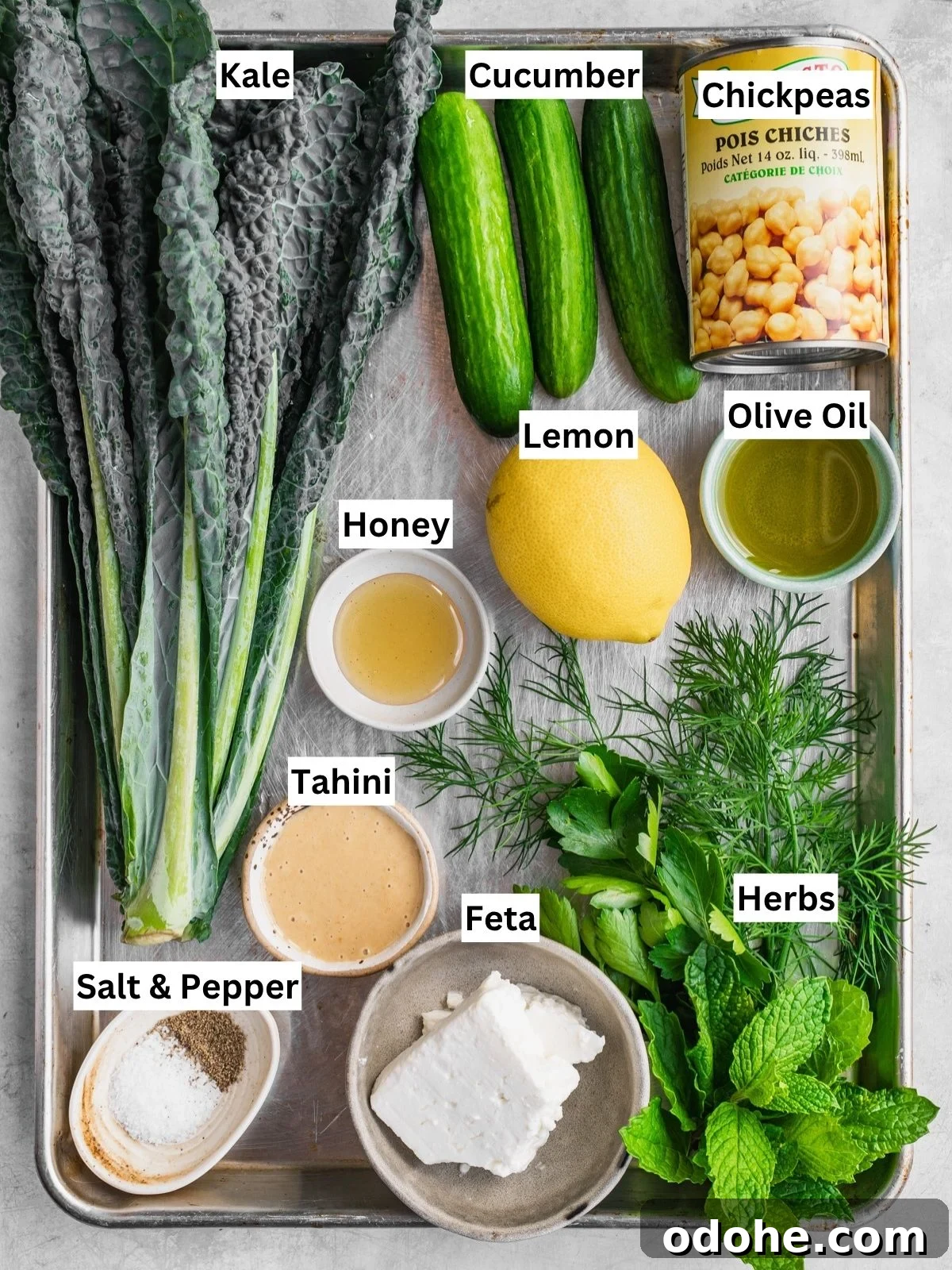 A tray with labeled ingredients for a Kale Chickpea Feta Salad: kale bunch, cucumbers, canned chickpeas, lemon, olive oil, honey, tahini, feta cheese, fresh herbs (dill and mint), plus small bowls of salt and pepper.