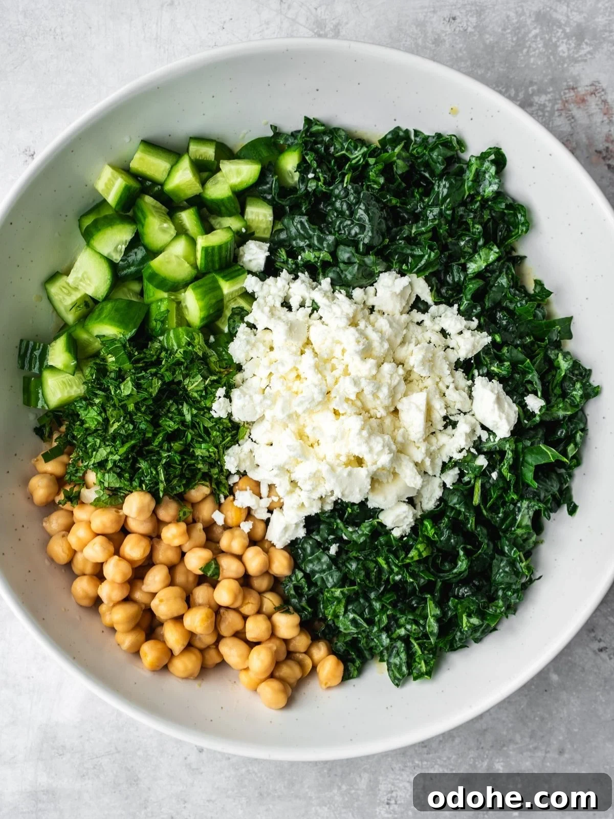 A bowl containing chopped cucumbers, kale, herbs, chickpeas, and crumbled feta cheese arranged in separate sections.