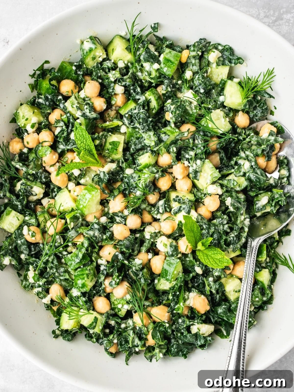 A white bowl of Kale Chickpea Feta Salad with chopped kale, chickpeas, cucumber, fresh mint, dill, and a creamy dressing, garnished with feta.