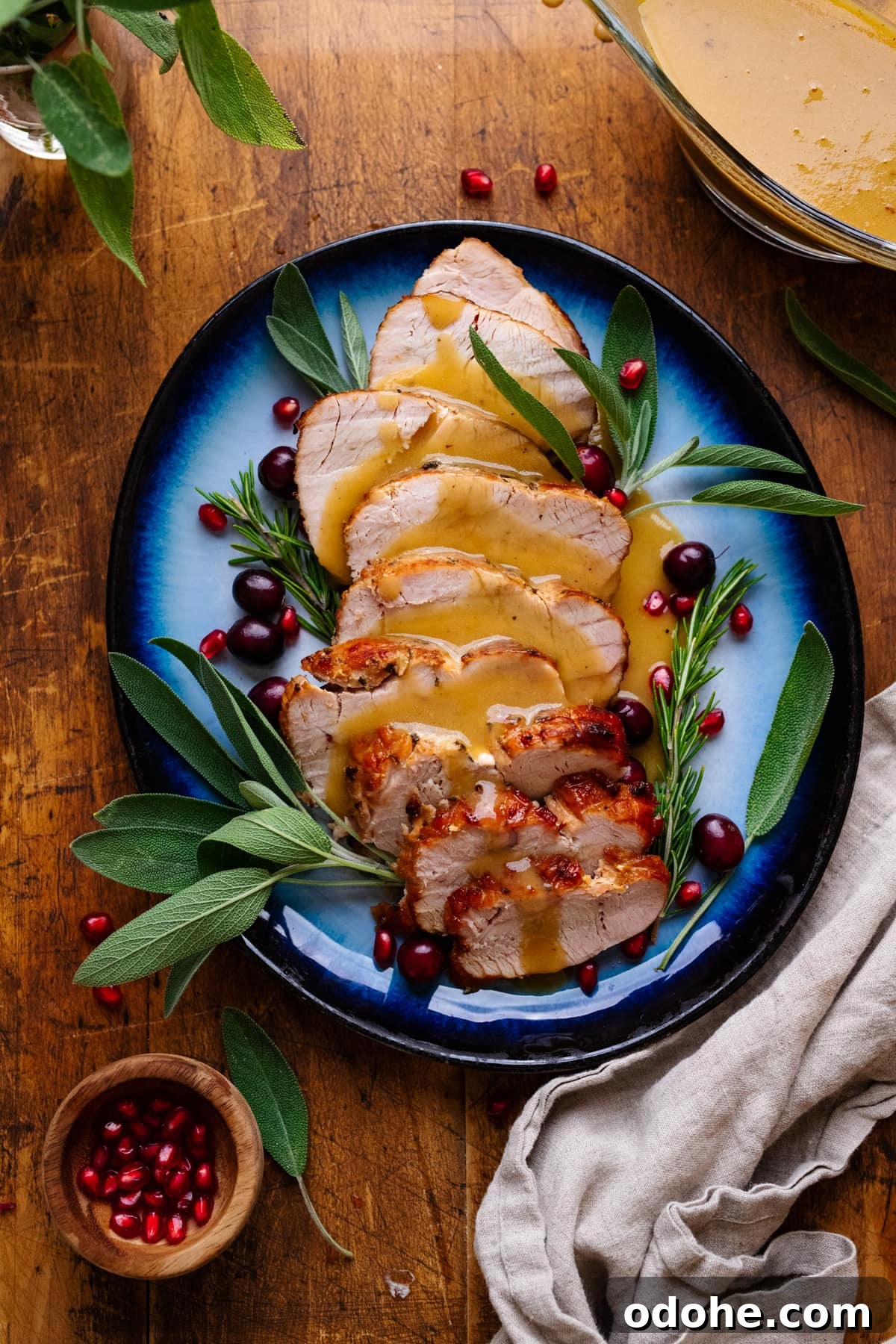 Maple Herb Turkey Breast in the Air Fryer: Juicy, Flavorful Roast 2 A blue plate with slices of air fryer turkey breast and gravy on it with fresh herbs arranged around it. The plate of turkey is arranged on a wooden background with herbs, gravy, and a gray napkin displayed around it.
