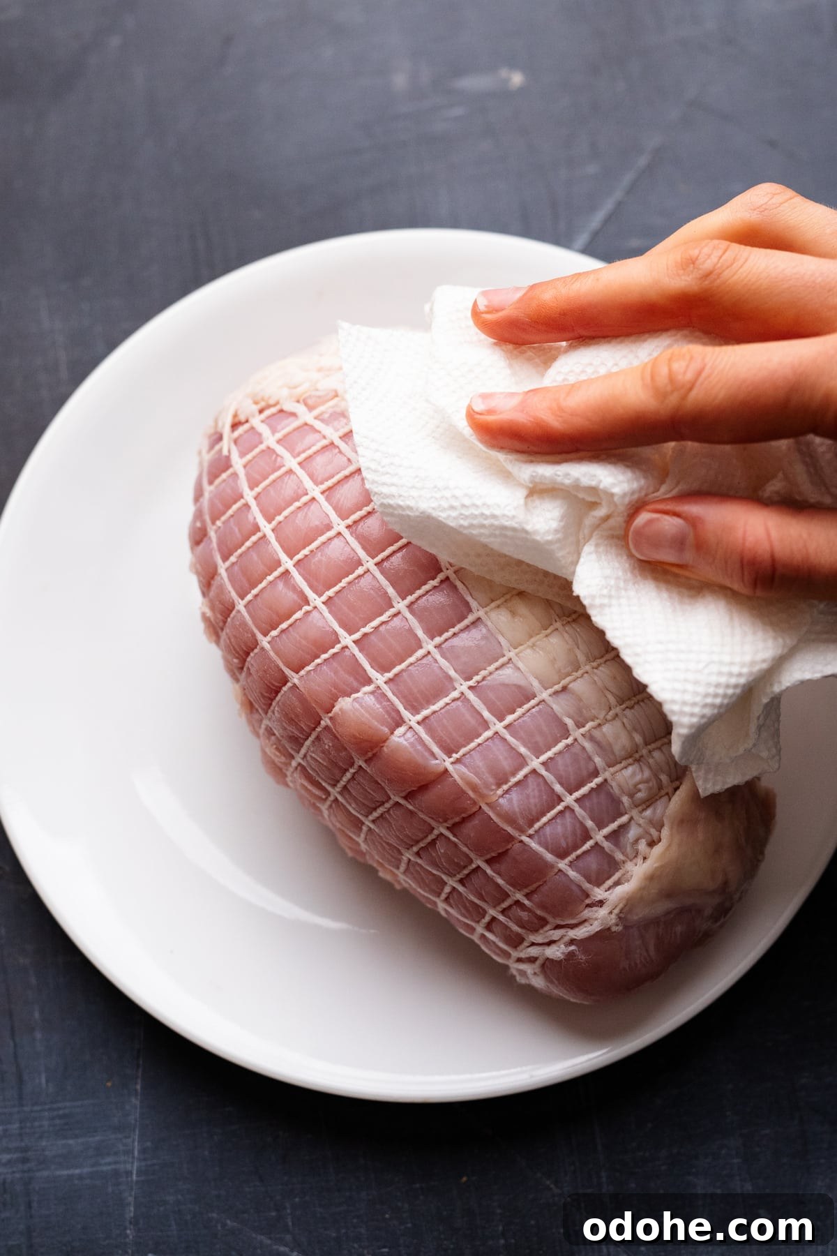 Maple Herb Turkey Breast in the Air Fryer: Juicy, Flavorful Roast 4 Hands patting paper towels on a turkey breast that's sitting on a white plate on a gray background.