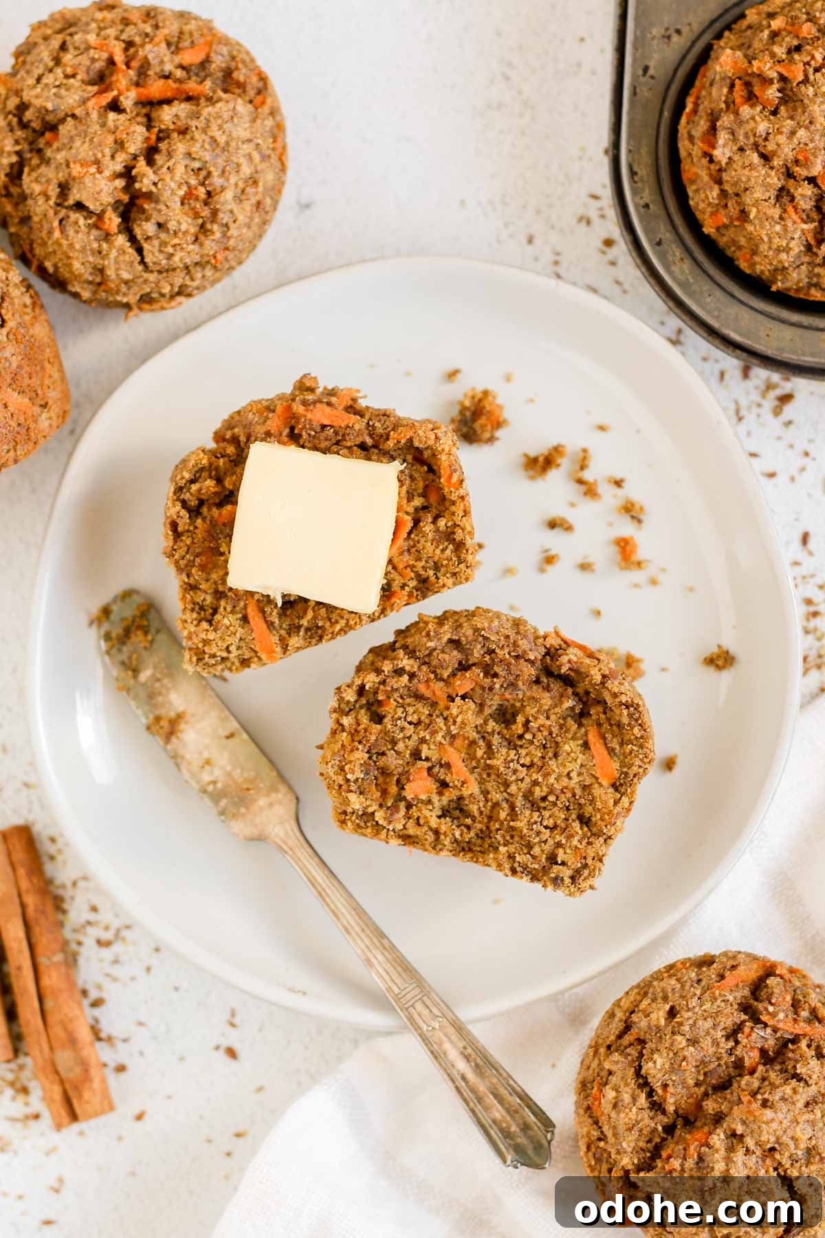 Flaxseed Carrot Muffins with Cinnamon and Honey 10 Round plate with a carrot flaxseed muffin cut in half with a square of butter on one half and a small knife resting on the plate with additional muffins on the side.