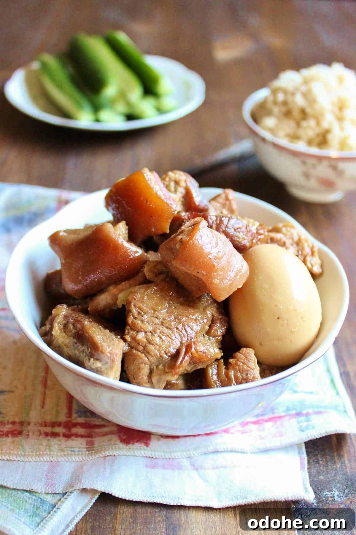 Vietnamese Braised Pork and Eggs (Thịt Kho Trứng) 2 Vietnamese Braised Pork with Egg in a bowl, a rice bowl is in the back along with a plate of cucumber.