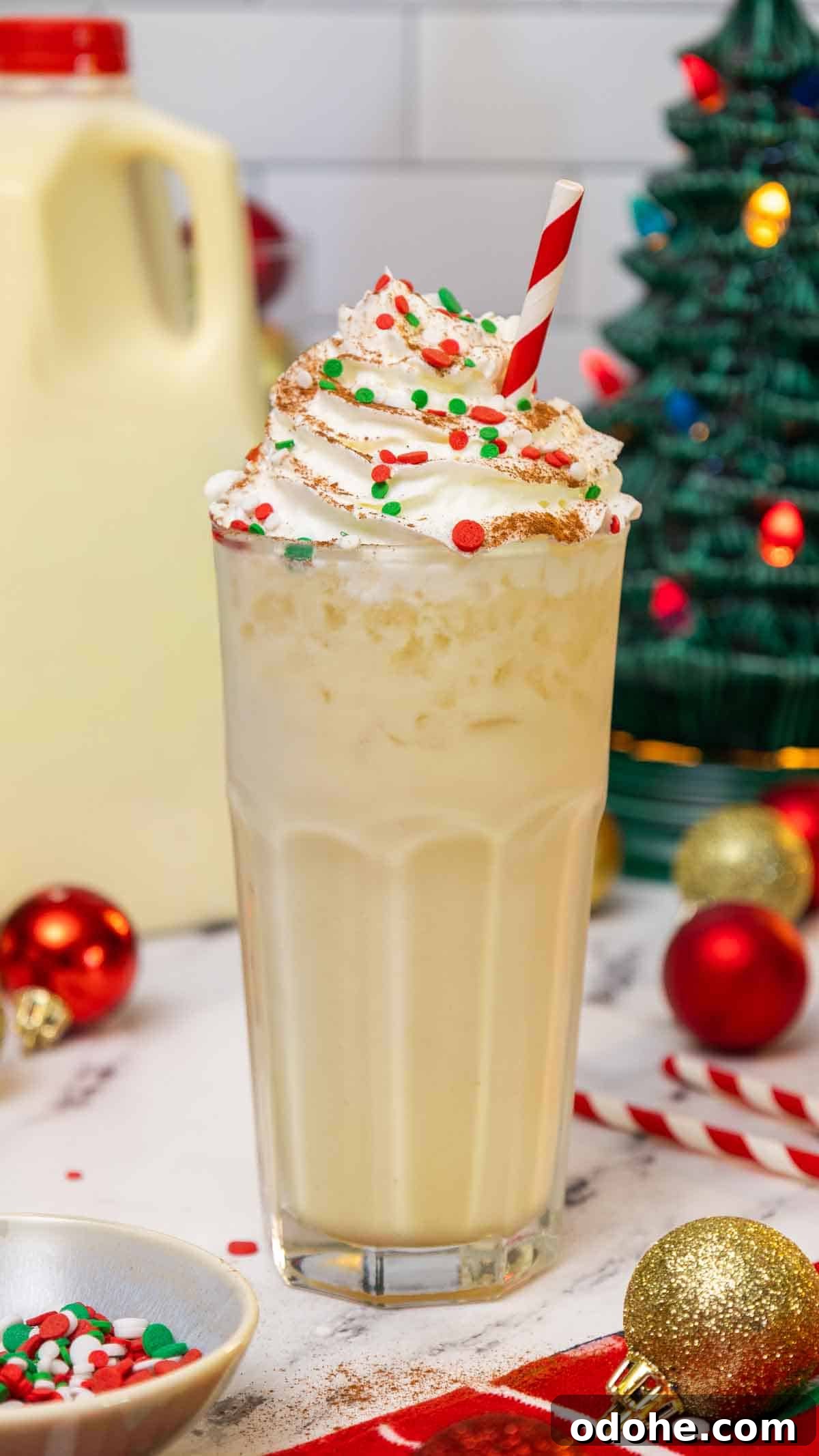 Tall glass of eggnog dirty soda topped with whipped cream, red and green holiday sprinkles, and a dusting of cinnamon, served with a red-striped straw. The festive background includes Christmas ornaments, a ceramic Christmas tree, and a carton of eggnog.