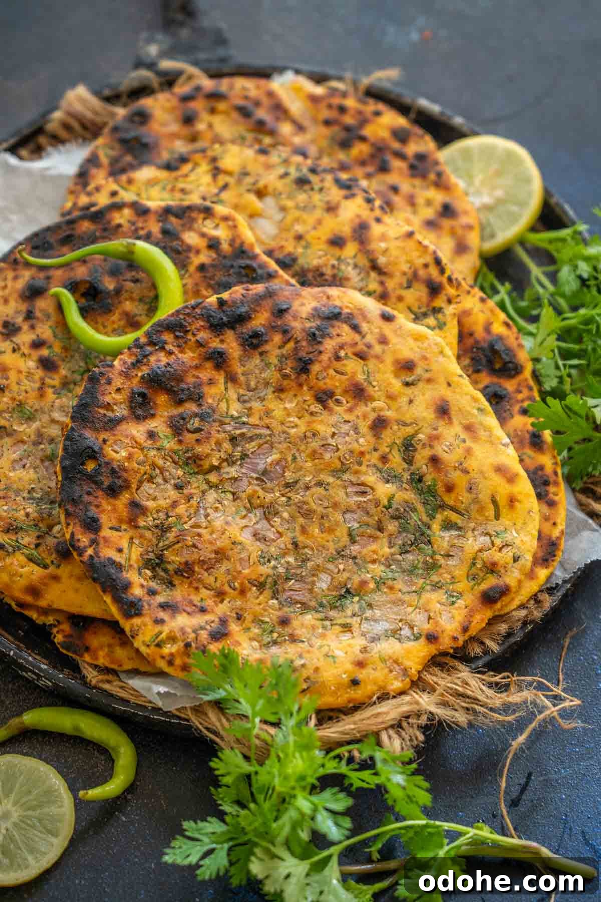 Three pieces of golden-brown, slightly charred Missi Roti garnished with herbs and onions, served on a rustic plate with green chili, cilantro, and lime slices on the side.