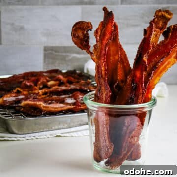 Crispy bacon strips in a clear glass jar, with more bacon cooling on a rack over a metal tray in the background.