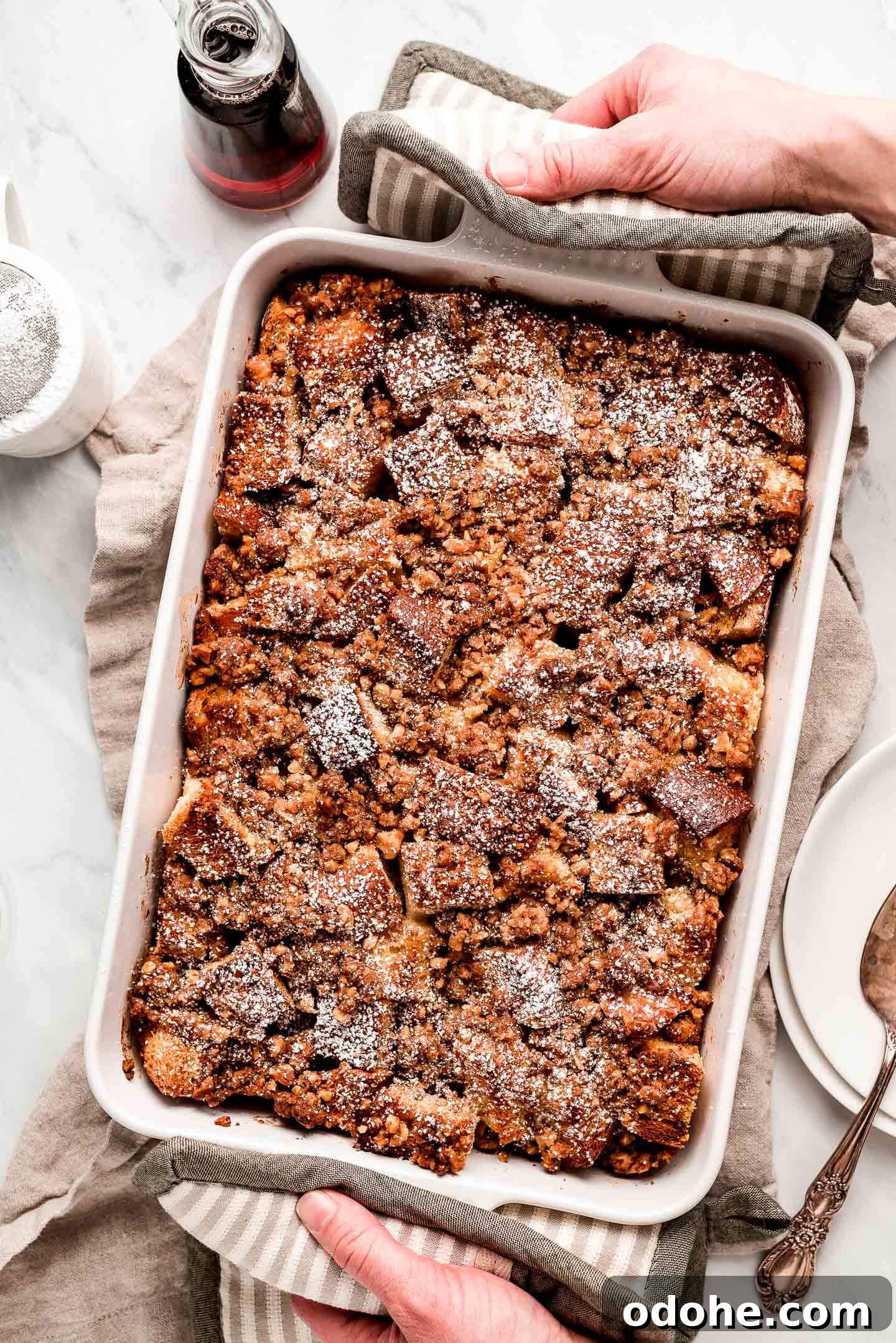 Make-Ahead Overnight French Toast Casserole Recipe 5 Hand with hot pads placing a pan of French Toast Bake on a marble surface with syrup, powdered sugar, and a piece of the casserole to the sides.