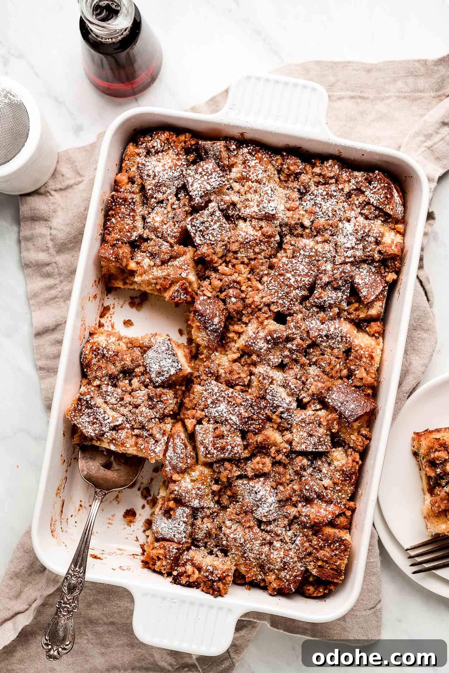 Make-Ahead Overnight French Toast Casserole Recipe 6 French Toast Bake on a marble surface with syrup, powdered sugar, and a piece of the casserole to the sides.