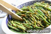 Asian style green beans in a white rimmed peacock patterned Polish pottery dish. A pair of wooden tongs are inserted into the dish on the left side.