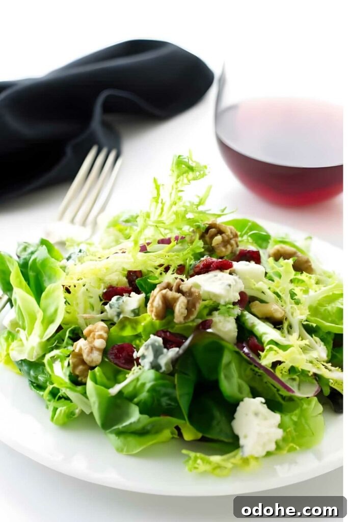 Green salad with walnuts, cranberries, gorgonzola cheese on white plate; wine behind.
