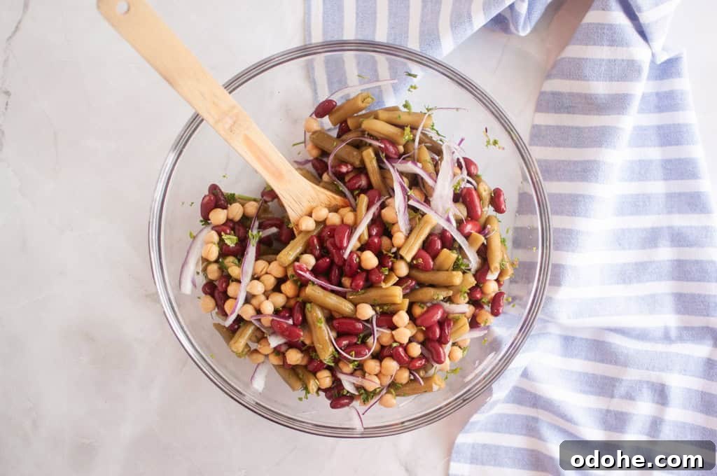 31 Memorial Day Recipes to Start Cookout Season Right 17 A glass bowl filled with a Three Bean Salad including kidney beans, chickpeas, green beans, and sliced red onion, with a wooden spoon and a striped cloth nearby.