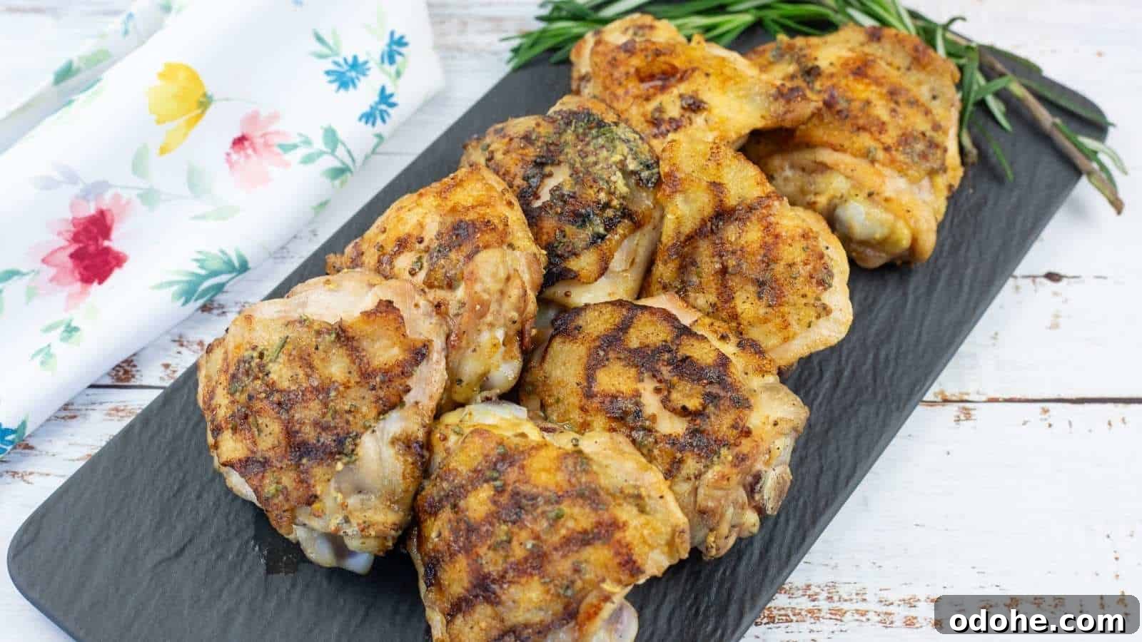31 Memorial Day Recipes to Start Cookout Season Right 20 Grilled Dijon rosemary chicken thighs are arranged on a rectangular black slate platter, garnished with fresh rosemary, next to a white floral napkin on a wooden surface.