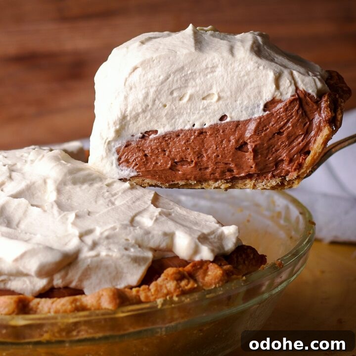 Someone using a spatula to lift a slice of chocolate cream pie from the whole pie so you can see the thick layers of chocolate cream and whipped cream.