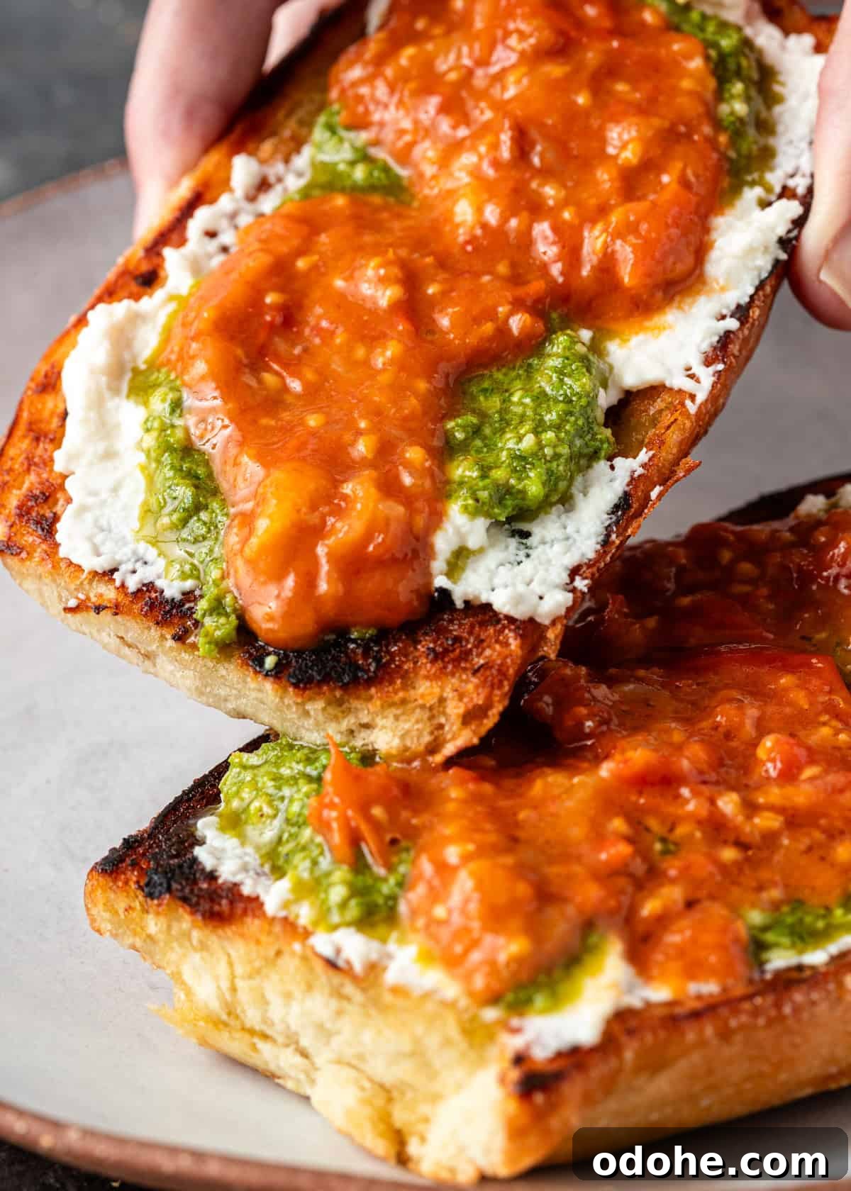 Slow-Roasted Cherry Tomato Confit Recipe for Flavorful Dishes 12 closeup: a hand lifting a piece of toasted bread with cheese, pesto, and tomato confit