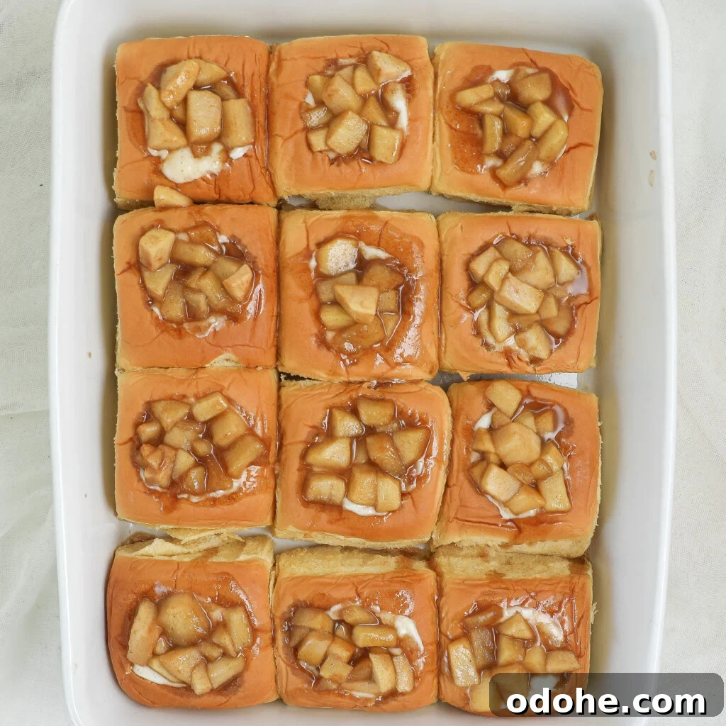 Twelve baked slider rolls in a white dish, each topped with a swirl of icing and a generous spoonful of caramelized diced apples.