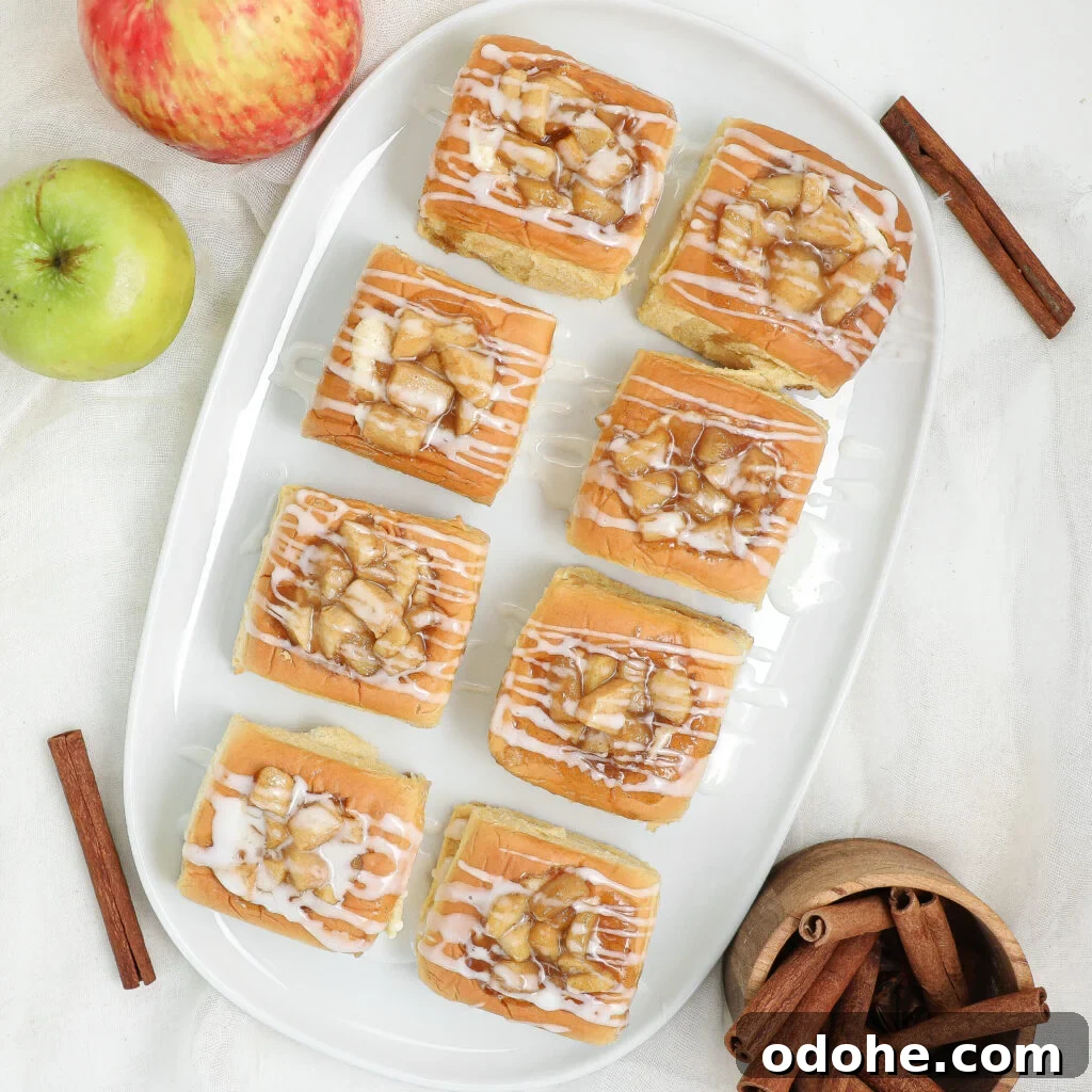A white platter holds nine square apple cinnamon rolls drizzled with white icing. Nearby are a red and a green apple, cinnamon sticks, and a wooden bowl of more cinnamon sticks on a light background.