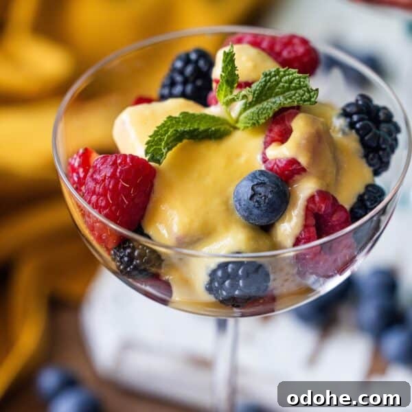 A glass dish filled with fresh raspberries, blackberries, and blueberries is topped with silky Berries and Creme Anglaise sauce and finished with a mint sprig.