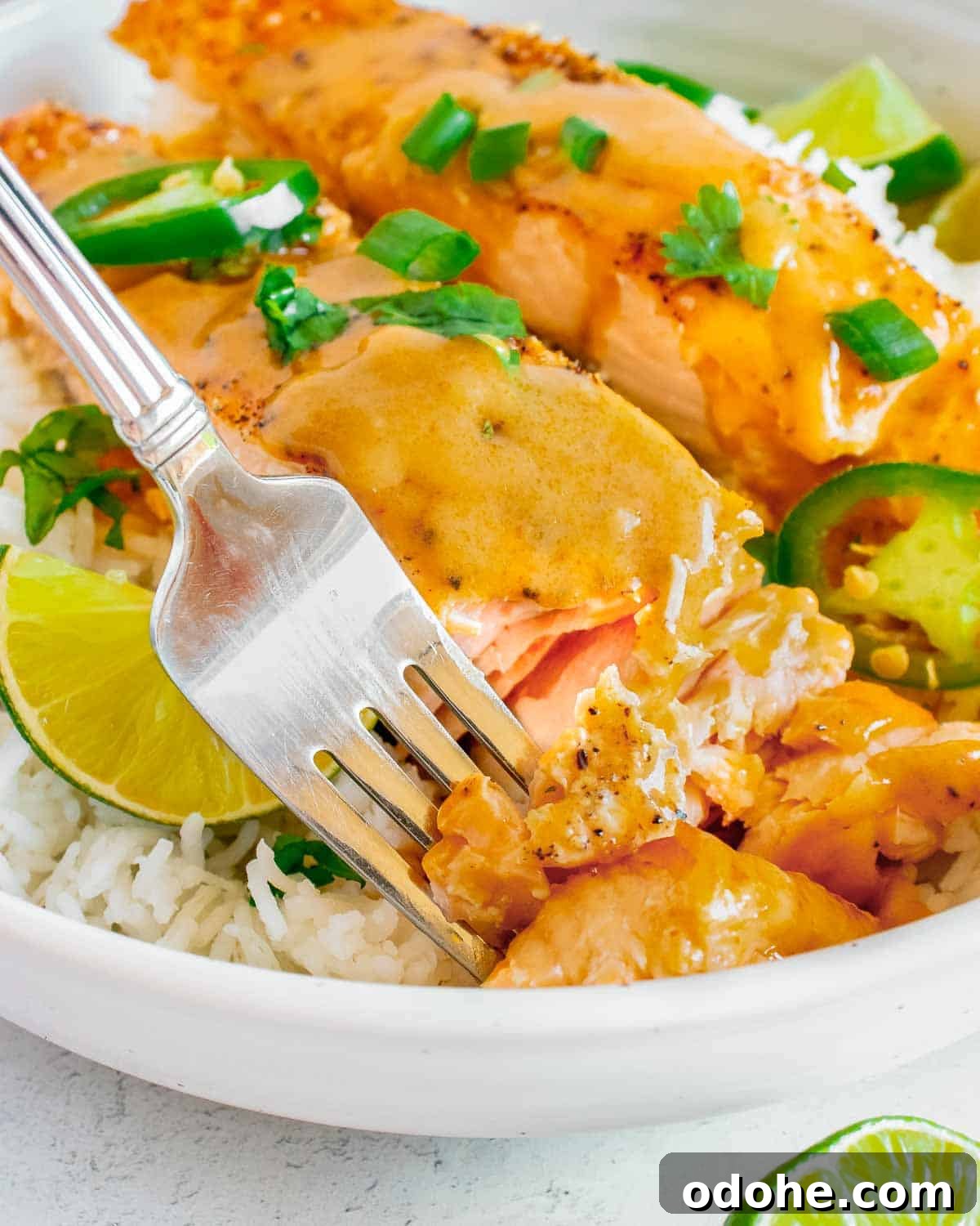 Thai-Style Salmon with Coconut Lime Sauce 6 Side close up shot of a fork picking up a piece of salmon garnished with parsley and sliced jalapeños on a bed of rice with lime wedges on the side of a large white bowl.