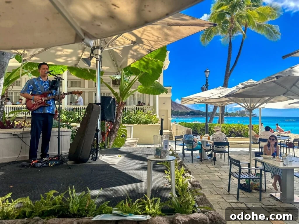 Musician performing with a guitar and singing at The Beach Bar of Moana Surfrider, a beachfront venue on Waikīkī Beach, with a backdrop of clear blue sky, ocean waves, swaying palm trees, and a glimpse of Diamond Head in the distance.