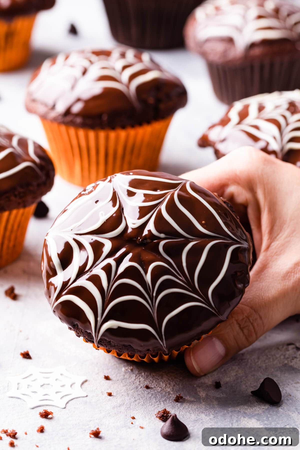 Chocolate Fudge Spiderweb Muffins Recipe — Decadent Halloween Treat 2 a hand holding a single chocolate fudge spiderweb muffin with some more muffins in the background.
