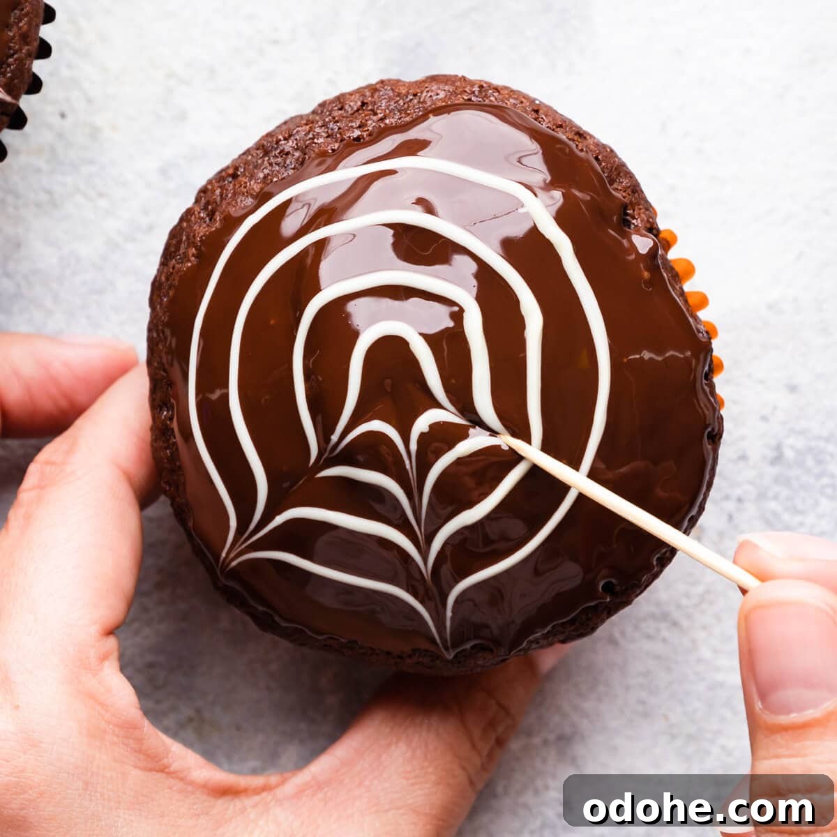 Chocolate Fudge Spiderweb Muffins Recipe — Decadent Halloween Treat 10 chocolate muffin being decorated with toothpick to create spiderweb design.