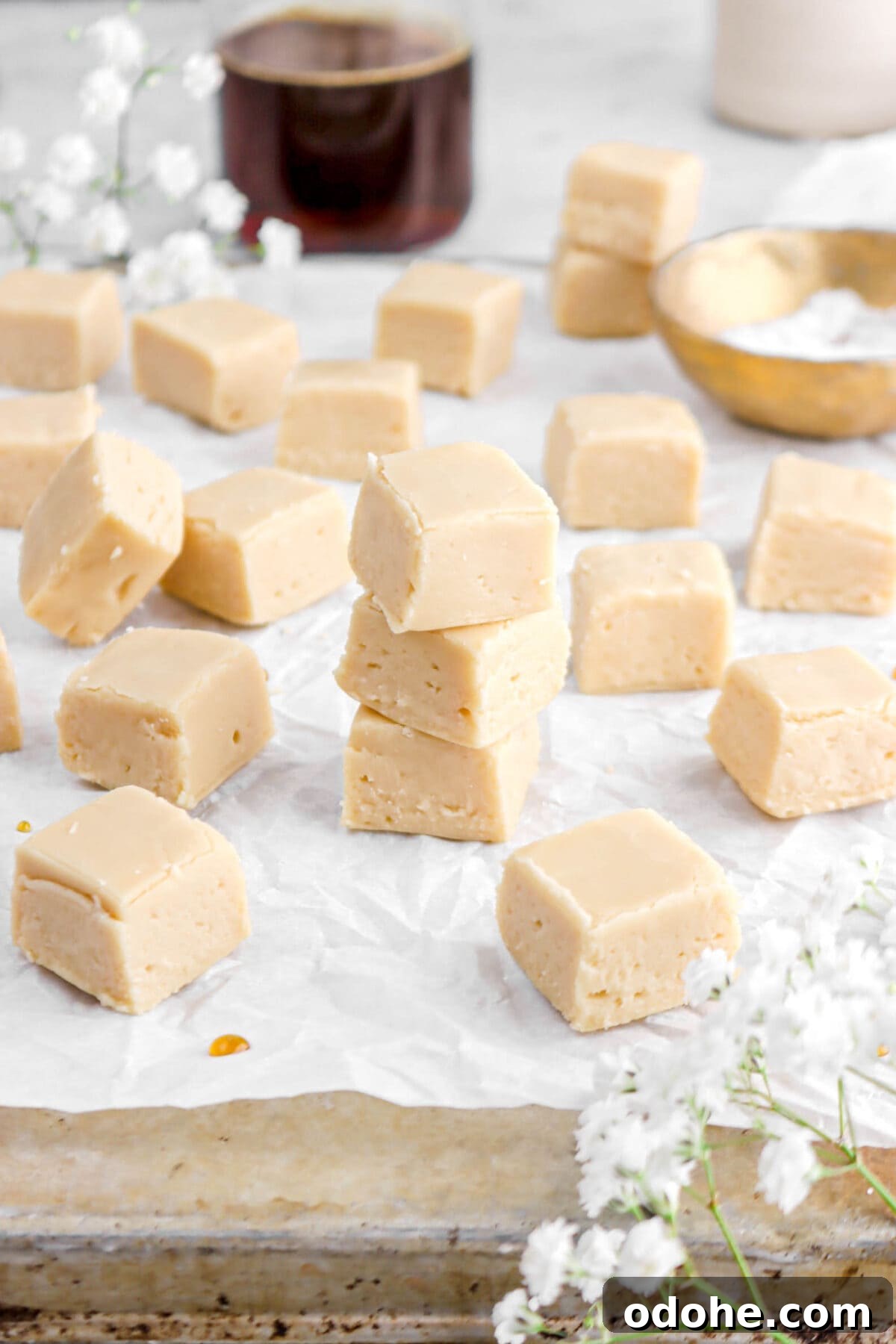 maple fudge on parchment paper on old sheet pan with white flowers around, gold bowl, and glass of maple syrup behind.