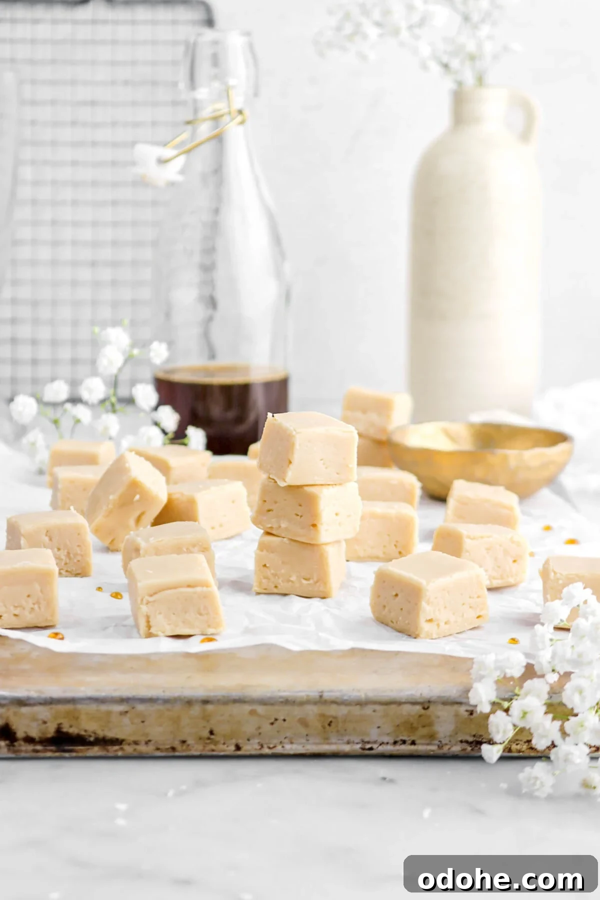maple fudge on upside down sheet pan with gold bowl behind, glass of maple syrup, and vase behind on marble surface.