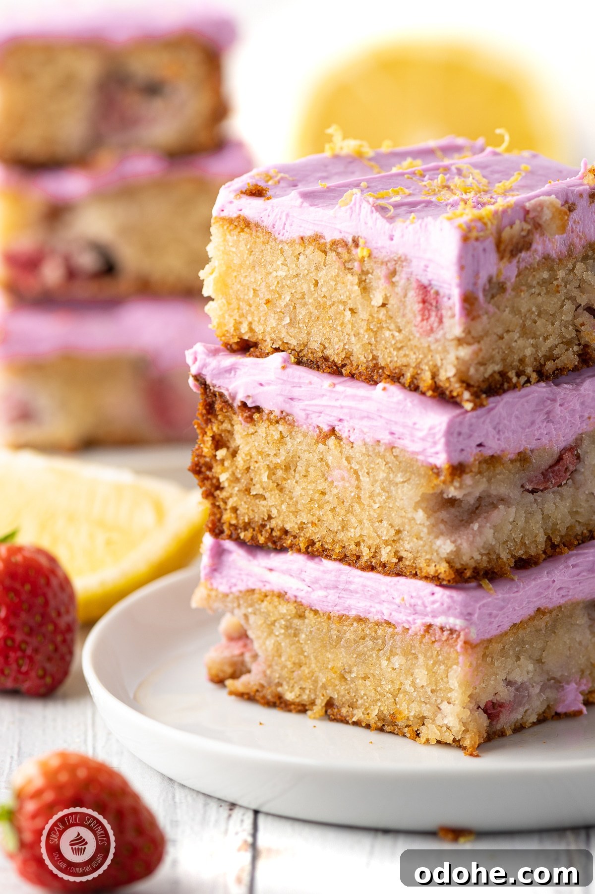 Three stacked low carb strawberry lemon blondies with pink frosting on a white plate with lemon wedges  and strawberries .