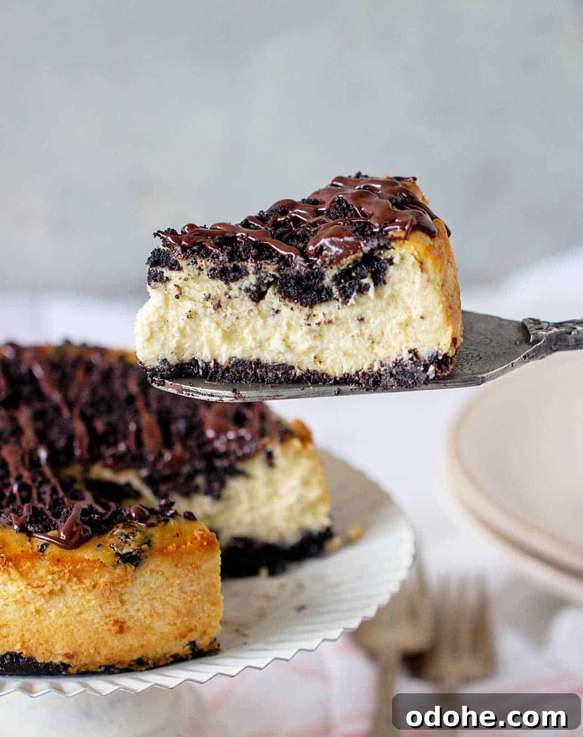 Whole oreo cheesecake on white cake stand with single slice floating above in silver cake server.