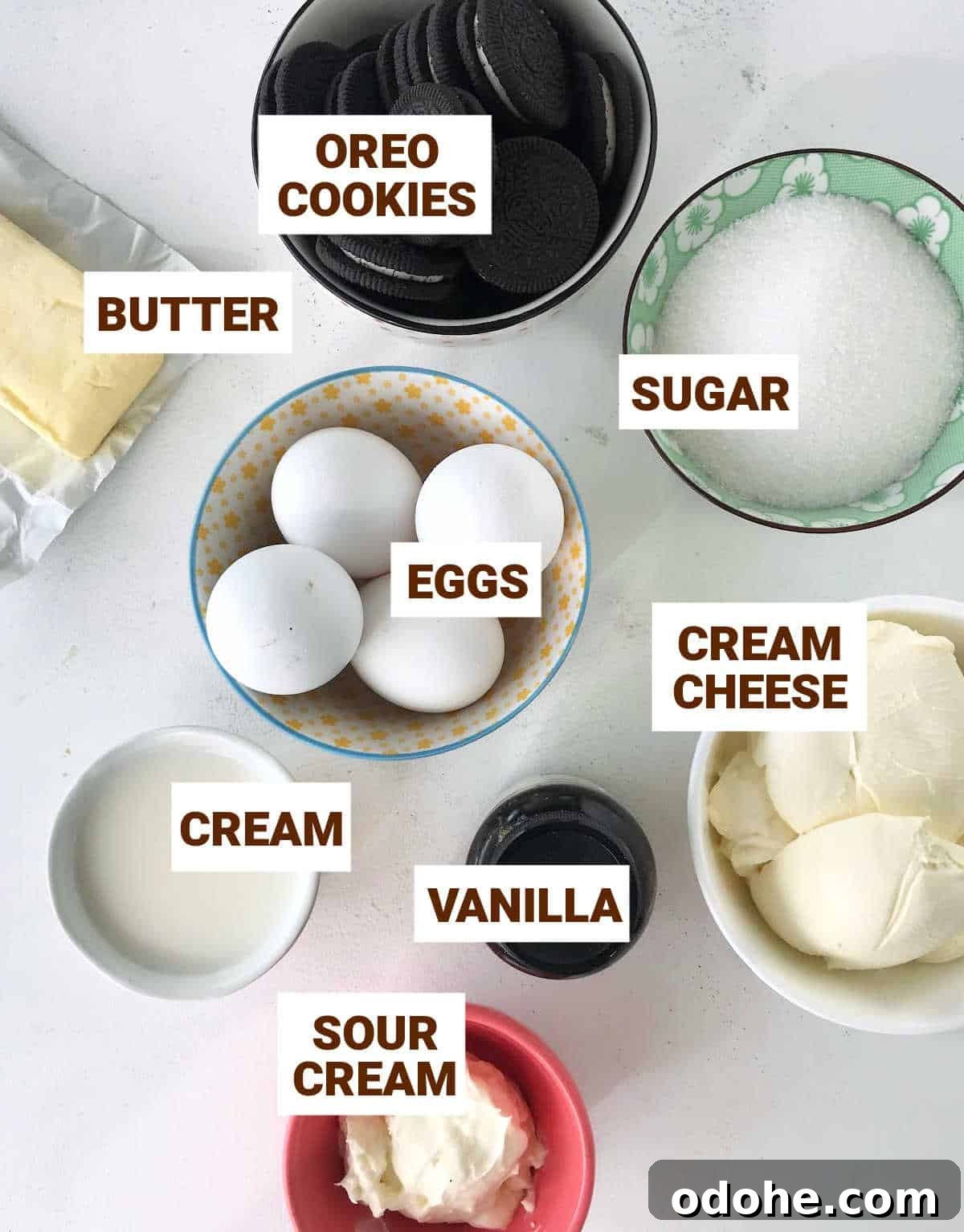 White surface with colorful bowl containing Oreo cheesecake ingredients such as eggs, cream cheese and butter.