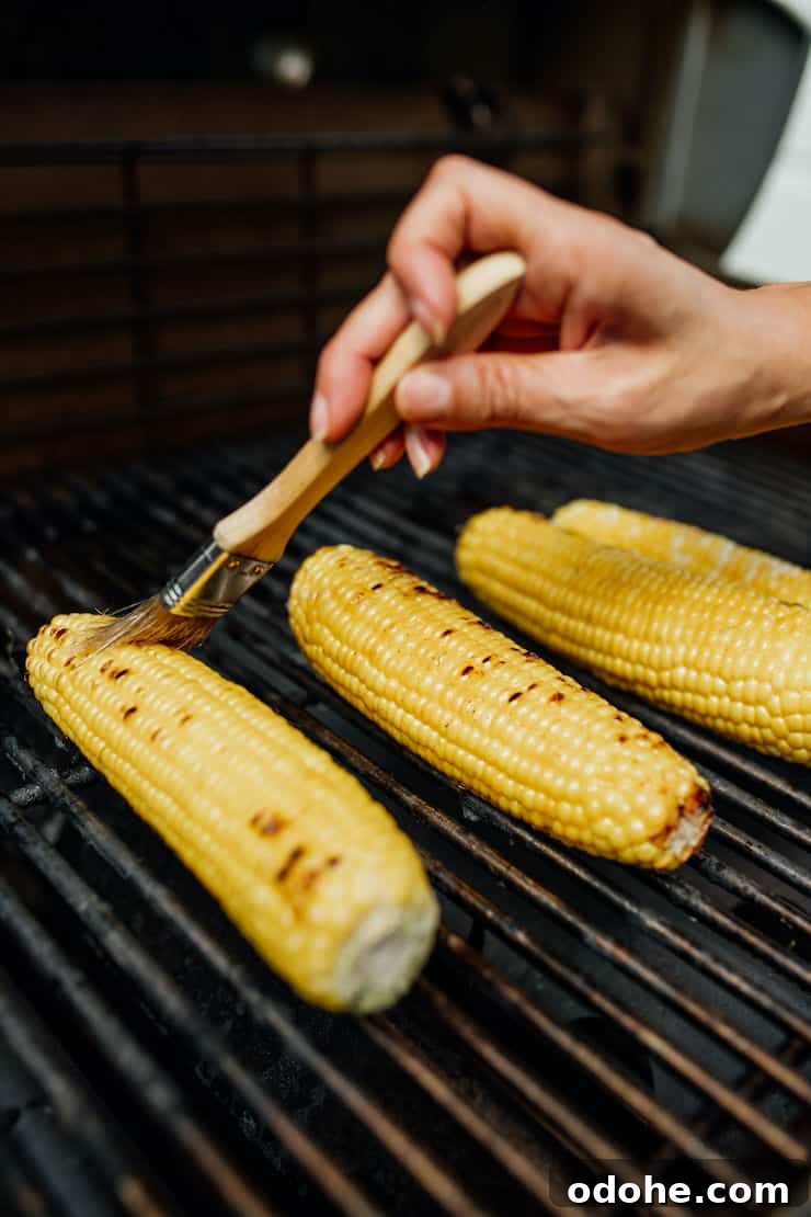 A hand brushing oil onto corn on the cob as it cooks on the grill, prepping it for Mexican Street Corn Salad.