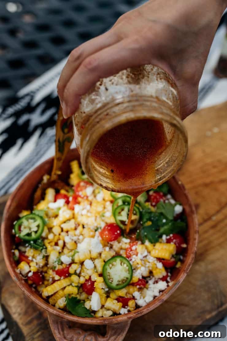A hand holding a mason jar and drizzling lime dressing over a bowl of Mexican Street Corn Salad with grilled corn, tomatoes, jalapeños, and Cotija cheese.