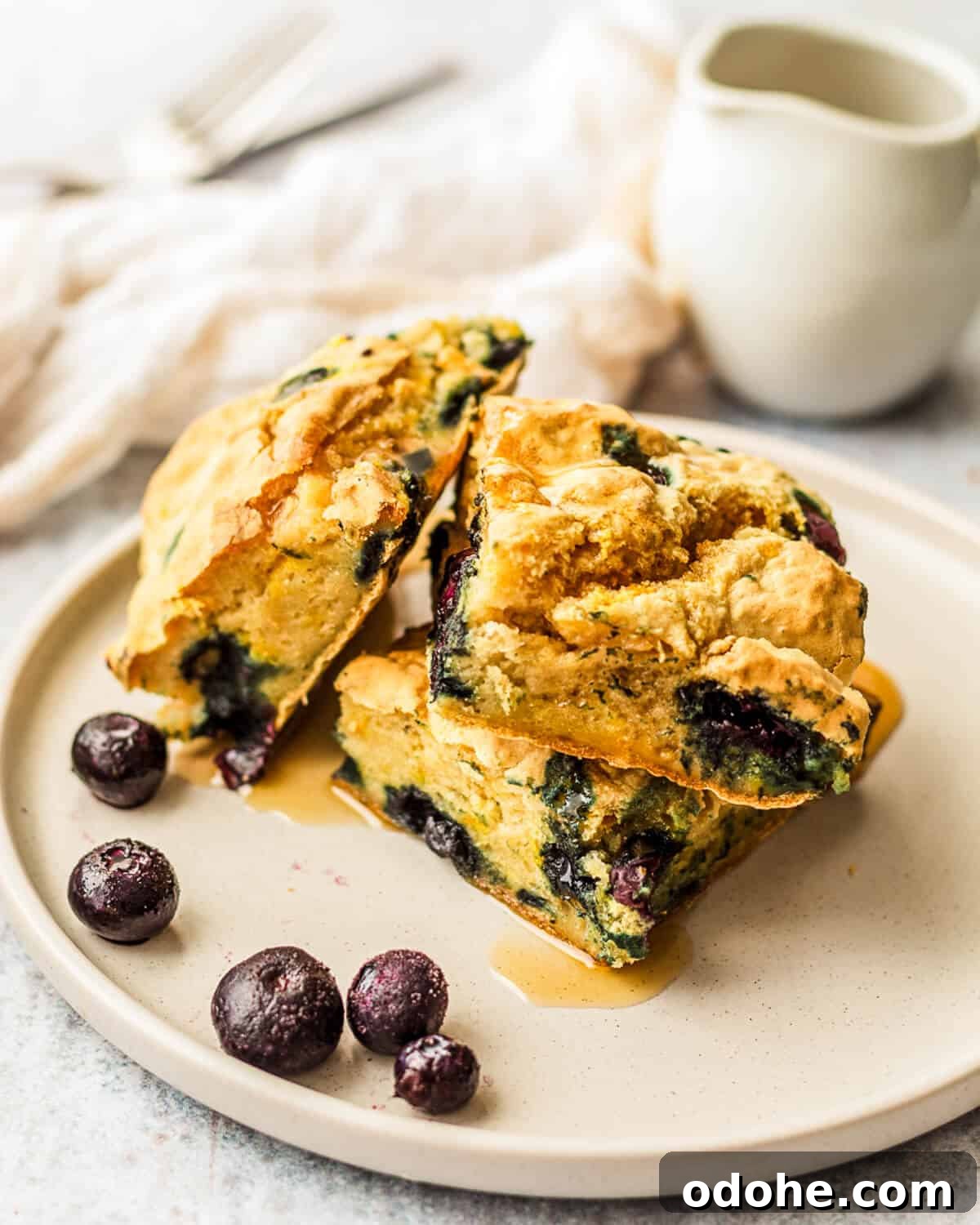 Three pieces of gluten-free sheet pan pancakes with blueberries and maple syrup poured over on a plate. There is a sauce jug in the background.
