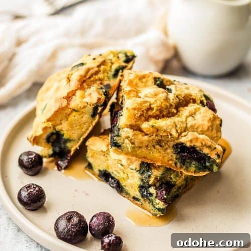 Three pieces of gluten-free sheet pan pancakes with blueberries and maple syrup poured over on a plate. There is a sauce jug in the background.