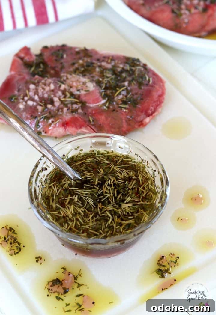 A clear glass bowl of marinade with a lamb steak on a white cutting board.