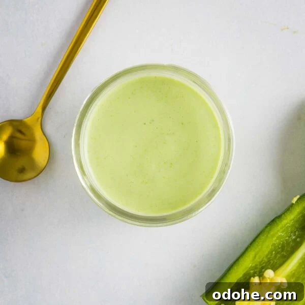 An overhead image of jalapeno ranch dressing in a glass bowl with a golden spoon on the upper left side and a jalapeno on the lower right side.