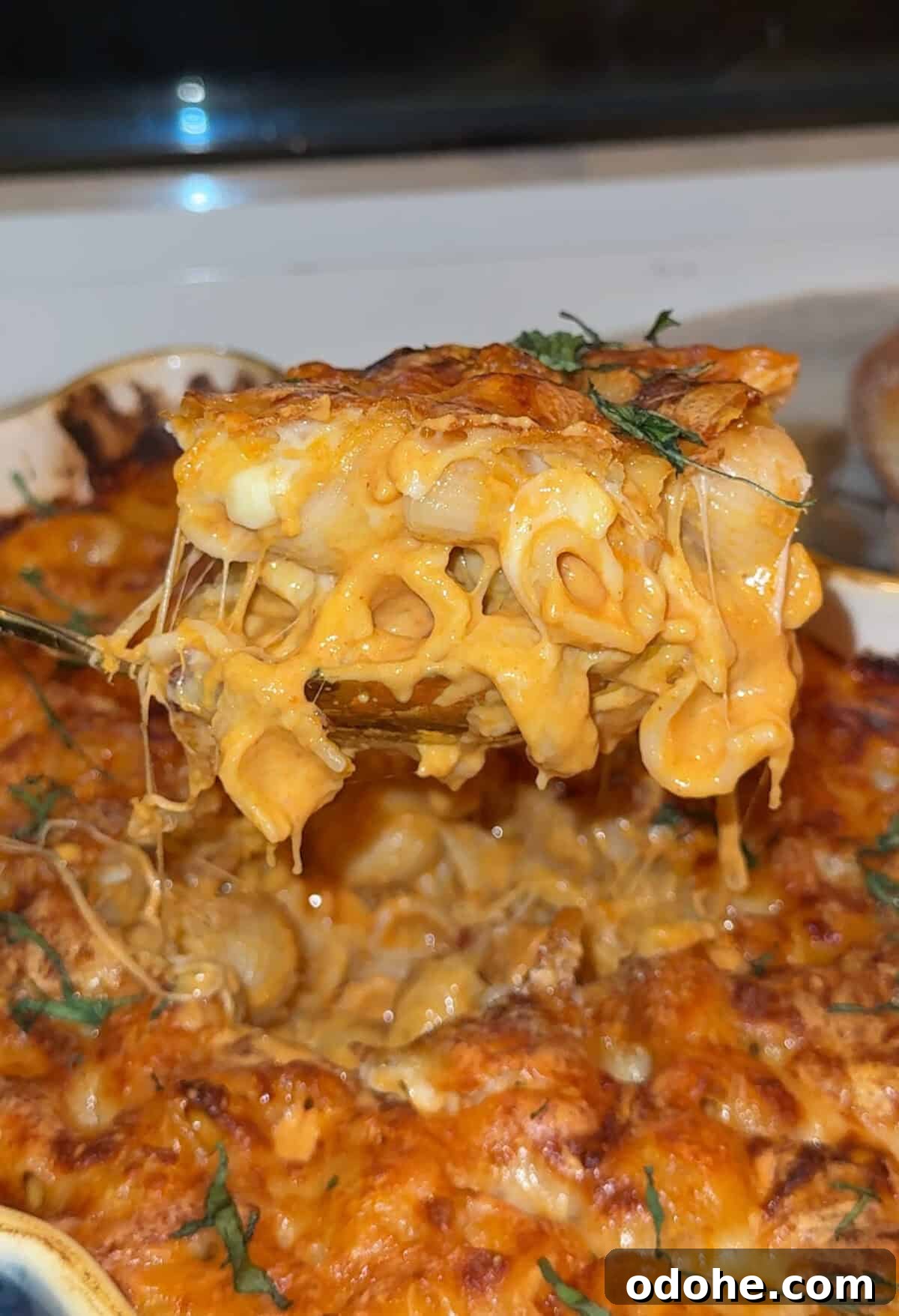 A close-up of a cheesy baked macaroni and cheese being lifted from a casserole dish, with gooey melted cheese stretching between the pasta and the pan.