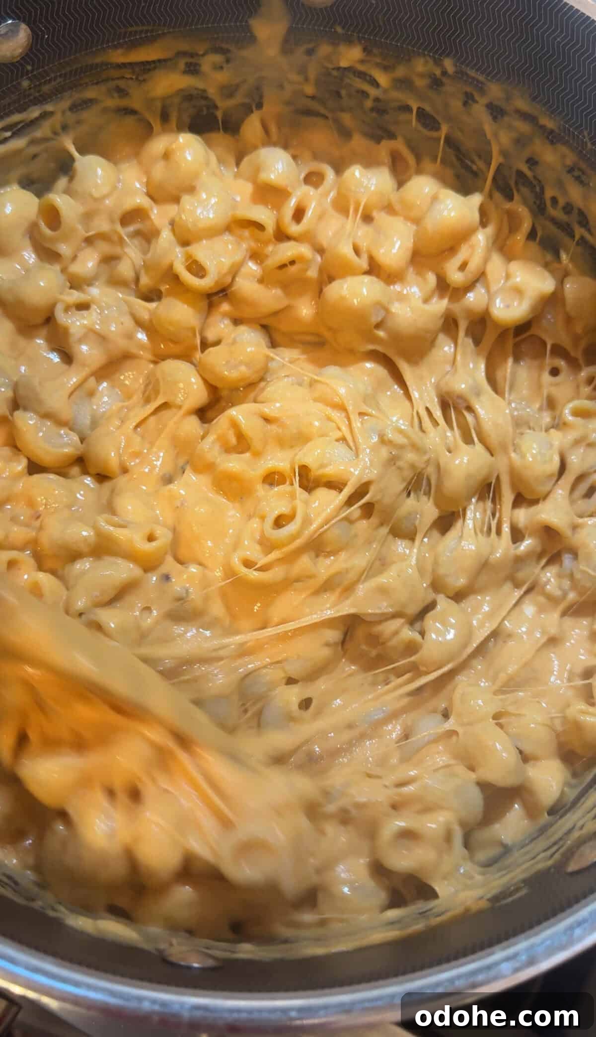 Close-up of creamy, cheesy macaroni shells being stirred in a pot, with melted cheese stretching between the pasta pieces.