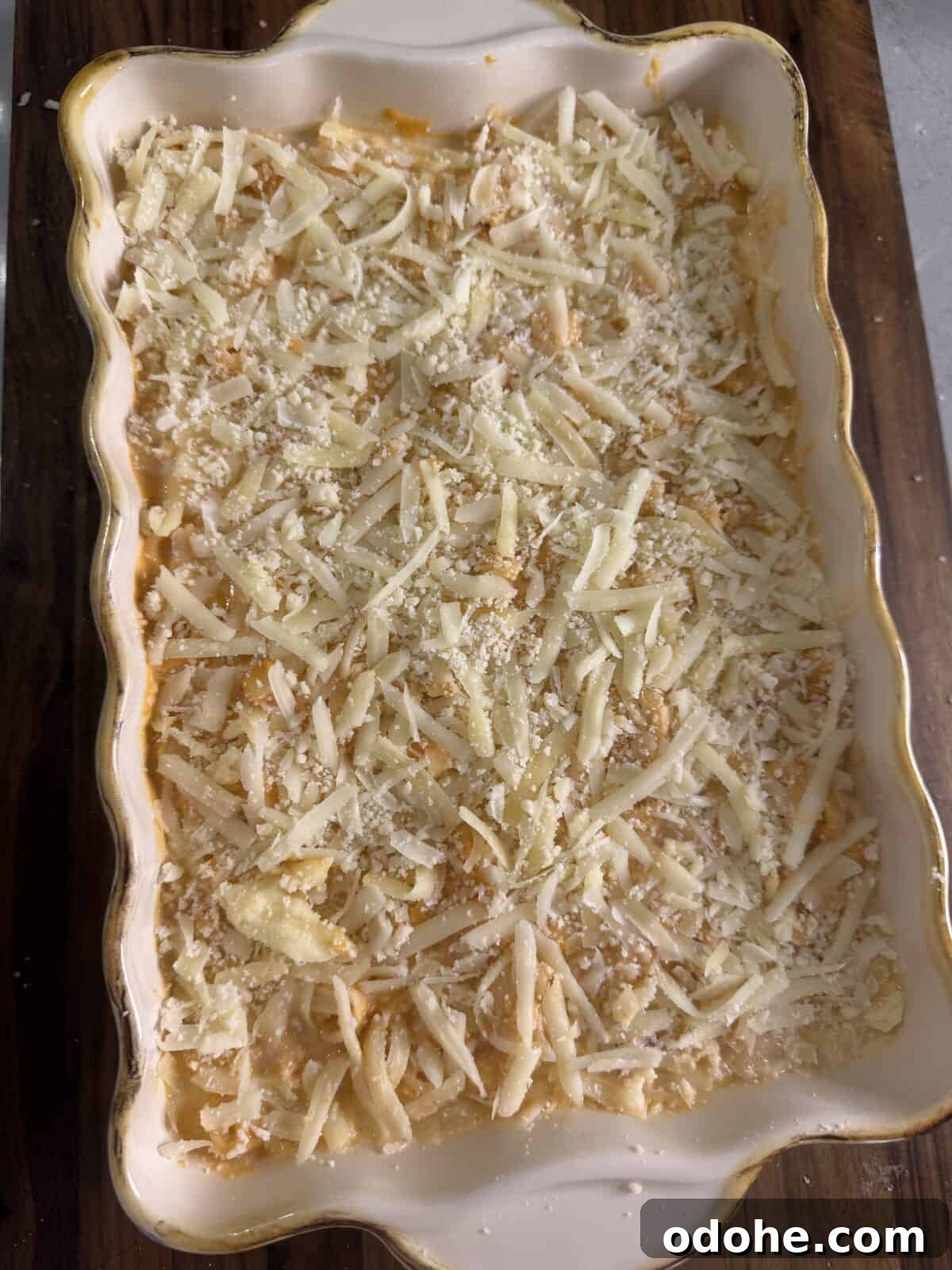 A rectangular ceramic baking dish filled with an unbaked casserole topped with shredded cheese and breadcrumbs, sitting on a wooden surface.