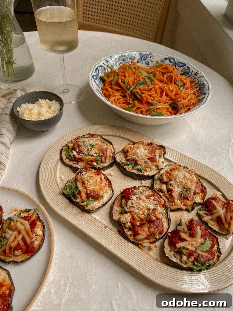 eggplant pizzas on a dish with a bowl of spaghetti and small bowl of parmesan and glass of white wine on a table